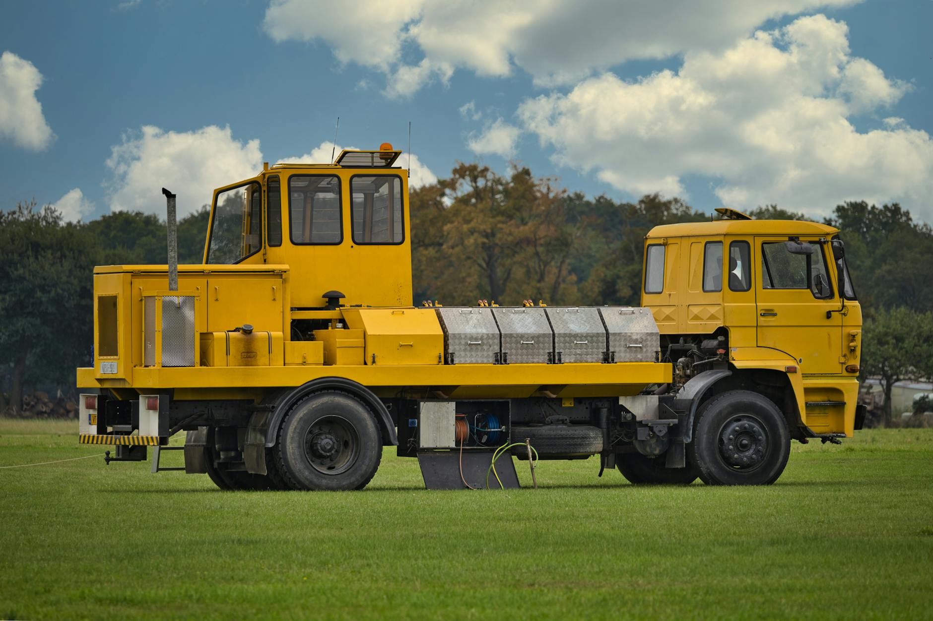 A yellow industrial vehicle parked on a lush green field under a clear blue sky with fluffy clouds.